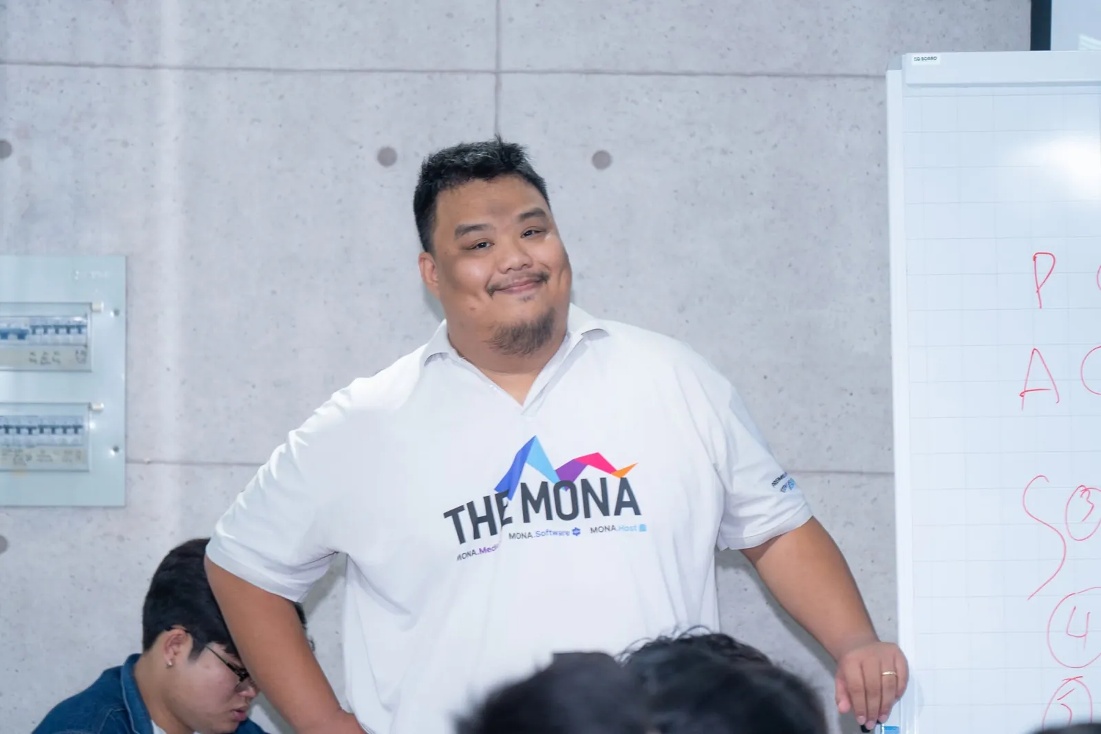 Khánh Hùng mặc áo THE MONA đứng lớp cạnh bảng trắng